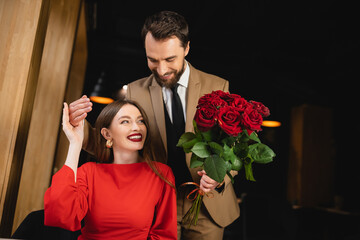 bearded man in formal wear holding bouquet of red roses near cheerful woman on valentines day