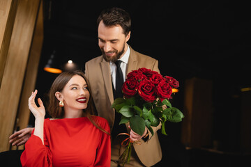 bearded man in formal wear holding bouquet of red roses near happy woman on valentines day