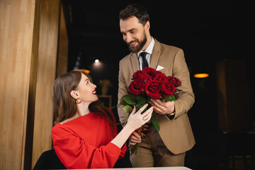 bearded man in formal wear holding bouquet of red roses near pleased woman on valentines day