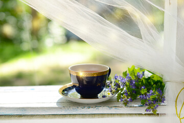 Cup of tea and blue flower bouquet on white wooden table on a terrace in summer morning. Herbal Tea, wildflowers and herbs on a blurred green garden trees bokeh background