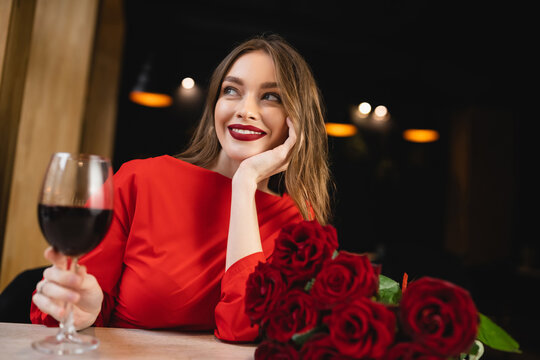 cheerful young woman holding glass of red wine near roses on valentines day