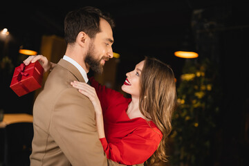 side view of cheerful woman holding present and hugging bearded boyfriend on valentines day