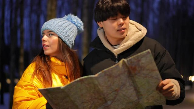 Lost Asian man and Caucasian woman with paper map standing outdoors at winter night talking looking around. Unsure couple of young tourists choosing route. Navigation and travelling