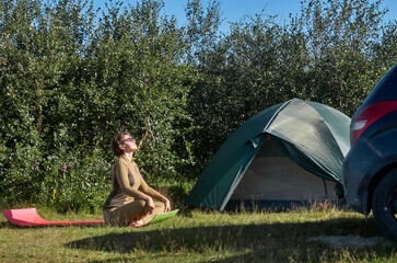 A female auto traveler performs yoga exercises in the open air. Nearby - tent and car