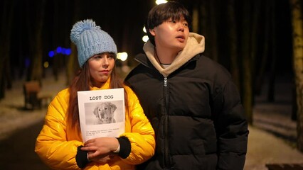 Handsome Asian man cheering up sad Caucasian woman standing with lost dog placard on night street on winter. Portrait of loving boyfriend endorsing girlfriend looking for pet outdoors in darkness