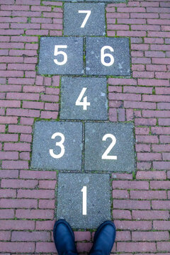 On The Paving Slabs Are Drawn Squares With Numbers - A Children's Game Of Hopscotch. Near The Boots Of An Adult Who Remembered Childhood.