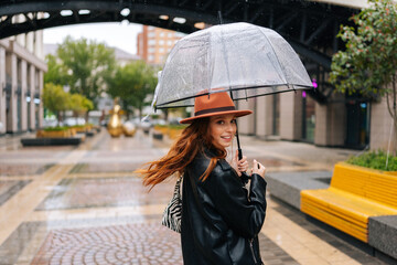 Back view of joyful redhead young woman in fashion hat walking on beautiful city street with transparent umbrella enjoying rainy weather outdoors, looking at camera. Concept of female lifestyle.