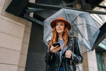 Low-angle view of charming redhead young woman wearing stylish hat using typing smartphone smiling looking away standing with transparent umbrella, by building in rainy European autumn city street.
