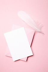 Holiday greeting card mockup with pink envelope and white feather on light pink background, top view, flat lay. Blank wedding invitation or Valentine Day letter, empty card