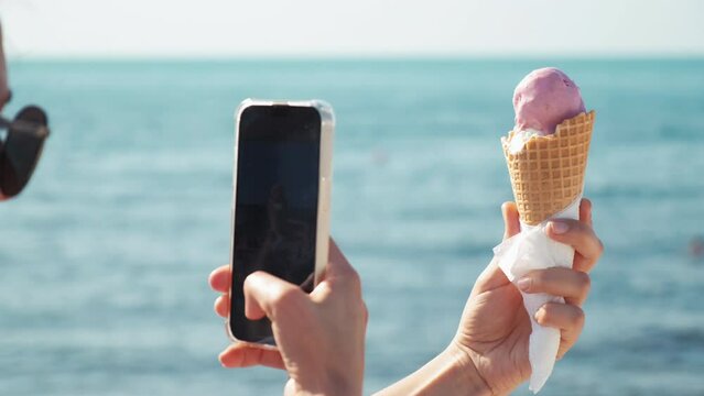 a girl takes pictures of ice cream on her phone on the beach. a girl eats ice cream in a cone on the beach. a girl goes with an ice cream cone on the beach. close-up of lips with strawberry ice cream