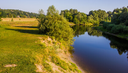 Fototapeta na wymiar a steep bank with a river cliff and slope with a tree covered with green grass and with trees and bushes and reflected in the water in a summer afternoon in sunny weather in Ukraine in Europe