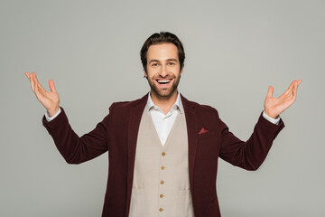 amazed showman in red blazer gesturing while looking at camera isolated on grey