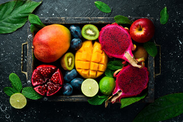 Tropical fruits in a wooden box: mango, dragon fruit, lime, pomegranate, plum, apple. On a black stone background.