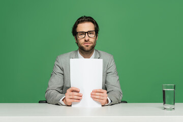 young broadcaster in eyeglasses and suit holding blank paper isolated on green