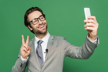 smiling reporter in eyeglasses and suit taking selfie on smartphone isolated on green