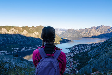 Hiking woman with backpack with scenic view of Kotor bay at sunrise in summer, Adriatic Mediterranean Sea, Montenegro, Balkan, Europe. Fjord winding along coastal towns. Sunbeams on Lovcen mountains