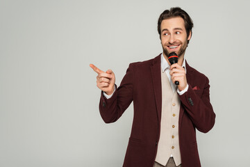 positive showman in formal wear talking in microphone and pointing with finger isolated on grey - obrazy, fototapety, plakaty