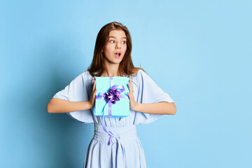 Portrait of young girl in cute tender dress posing with present box over blue background. Shocked expression. Gifts