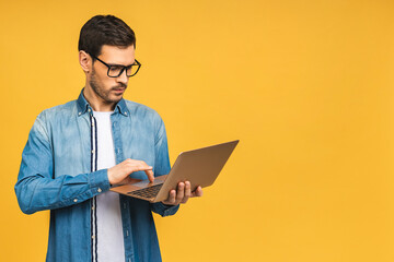 Confident business expert. Confident young handsome bearded man in casual holding laptop and smiling while standing over isolated yellow background. - 555902980