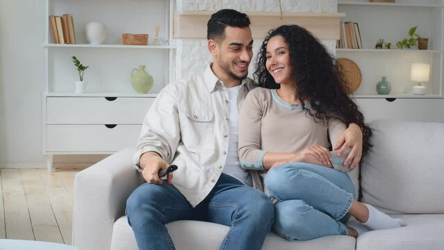 Multiethnic couple sitting at home watching movie film talking discussing. Hispanic Indian man husband with remote control and Caucasian woman wife hugging sit on sofa laughing discuss TV program