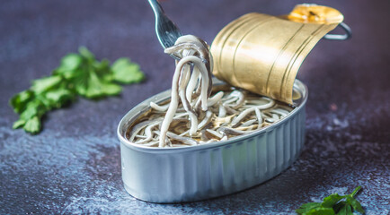 Fork pricking canned baby eels on a dark blue table with parsley. Eating preserve cans.