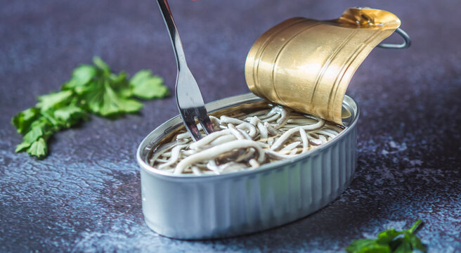 Fork pricking canned baby eels on a dark blue table with parsley. Eating preserve cans.