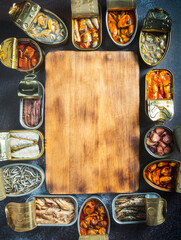 Preserve cans with different products, fish and seafood, natural or pickled, open on a dark table with a wooden cutting board and lemon. Ready to eat.