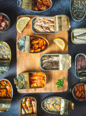 Preserve cans with different products, fish and seafood, natural or pickled, open on a dark table with a wooden cutting board and lemon. Ready to eat.