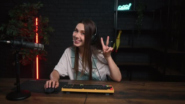 A cute streamer girl is playing online games on the computer and showing a peace gesture to the camera with a smile on her face.