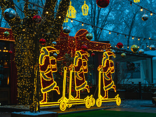 Festive lighting in the evening in the park. Lamps in the form of Santa Claus with a gift on a scooter. New Year's lighting.