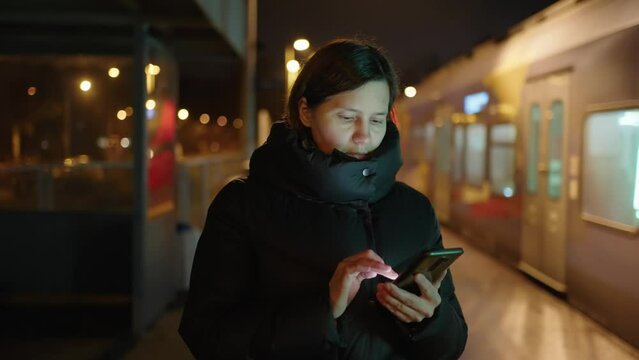 Woman in a black jacket uses a phone on the platform of a city train at night