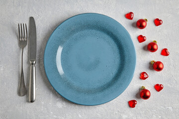 An empty blue ceramic plate with cutlery surrounded by red christmas balls and hearts on a light concrete table