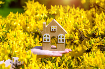 The girl holds the house symbol against the background of blossoming yellow rhododendron
