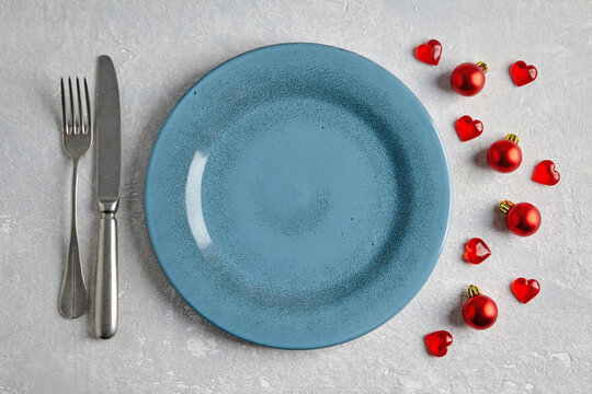 An empty blue ceramic plate with cutlery surrounded by red christmas balls and hearts on a light concrete table