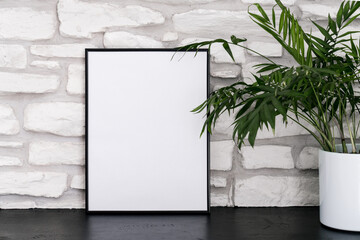 Mock-up of black vertical frame with white background. Green plant and empty poster on table. Shelf...