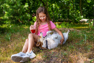 A beautiful and sweet girl sits on the grass with her beloved dog and drinks juice. A happy and smiling child walks in nature with his beloved pet in the summer in the park and drinks soda.