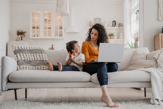 Overloaded  young Italian woman with curly hair dressed in orange blouse and dark blue pants sitting on sofa using laptop, working at home at the kitchen with son playing on phone, remote working.