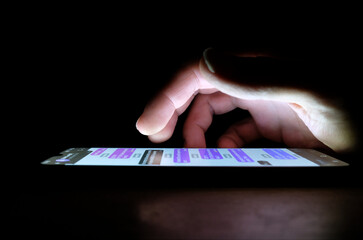 Hand using an app on a smartphone in the dark late at night with lighting from phone screen. Person...