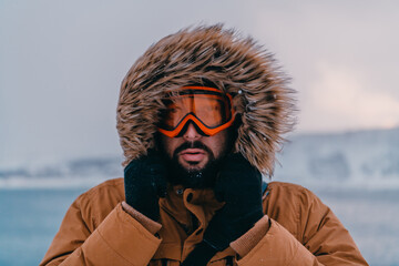 Fototapeta na wymiar Headshot photo of a man in a cold snowy area wearing a thick brown winter jacket, snow goggles and gloves. Life in cold regions of the country.