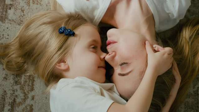 Top view from above faces of Caucasian mother and small little kid child lying on floor indoors talk speaking. Close up heads mom talking to baby kiss nose kissing girl play bite laugh touching noses