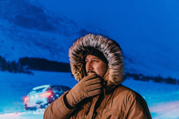 Fototapeta na wymiar Head shot of a man in a cold snowy area wearing a thick brown winter jacket, snow goggles and gloves on a cold Scandinavian night. Life in the cold regions of the country.
