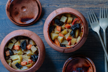 Stewed potato, carrot, onion, tomato and prunes in a clay pots with lid on wooden background, closeup