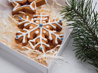 Christmas gingerbread cookies lie in a white box with straw near a pine branch. christmas cookies with icing. cookie in the form of a snowflake with blue dots. gingerbread lies on a white table.