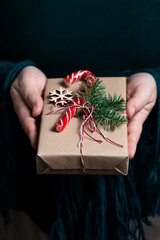 A woman holds in her hands a gift tied with a thread, decorated with a sprig of nobilis and a caramel stick.