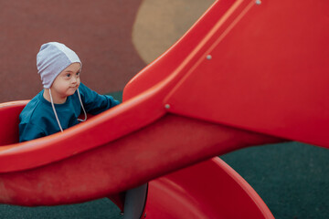 Cute Little boy with down syndrome in a funny hat walks in the playground, rolling down the...