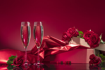 Luxurious glasses with sparkling wine open box with glitter brocade surrounded by roses on a mirror surface on a red background. Valentine's day or romantic evening invitation
