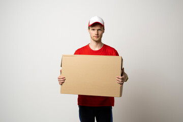 Portrait of a young bearded delivery man in red uniform holding blank cardboard, percel isolated on white background.