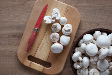 Fresh champignons on the table, in a bowl and on a wooden board with a red knife, top view of vegetable ingredients for cooking.