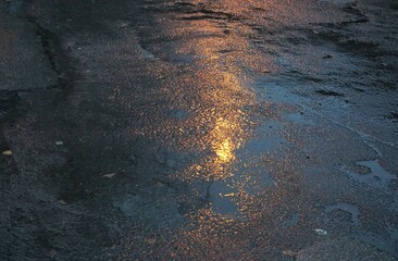 Alte nasse asphaltierte Straße mit Regenpfützen und goldener Lichtspiegelung bei Regen am Abend...