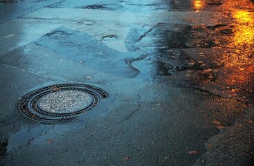 Alte marode nasse Straße mit Wasserpfützen und goldener Lichtspiegelung bei Regen am Abend im...